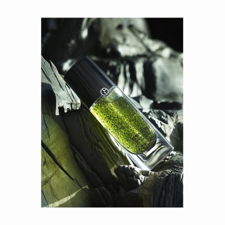 A glass perfume bottle with a black cap filled with green liquid sits on a jagged rock surface, set against a blurred rock backdrop.