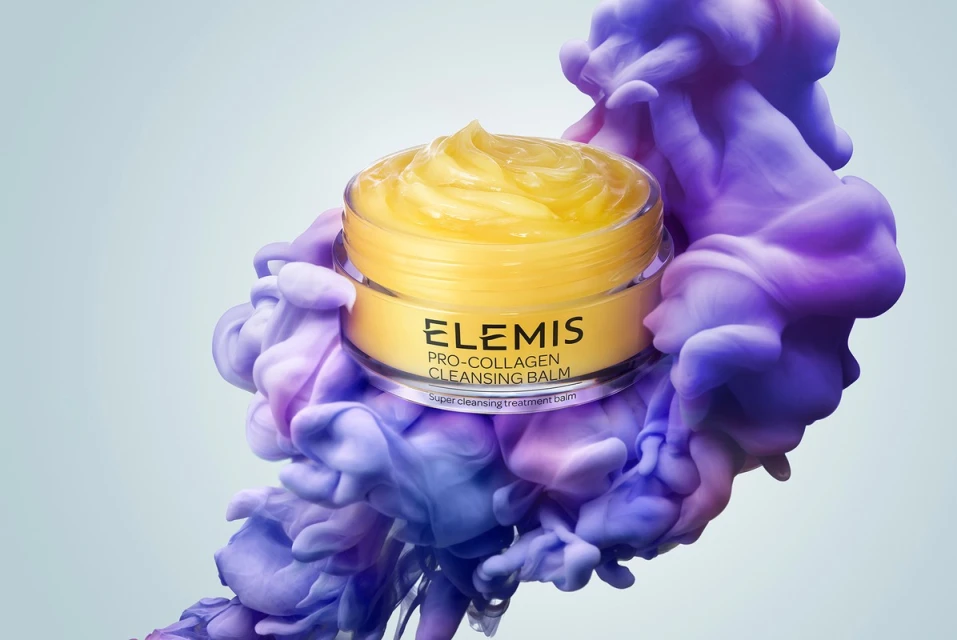 Close-up of Elemis Pro-Collagen Cleansing Balm jar, showcasing its golden contents and brand label, set against a background of swirling purple.