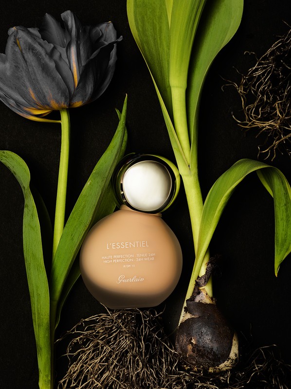 Still life shot of Guerlain L'Essential foundation bottle surrounded by black tulip and green plant stems against a black background.