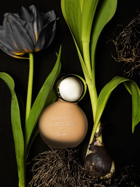 Still life shot of Guerlain L'Essential foundation bottle surrounded by black tulip and green plant stems against a black background.