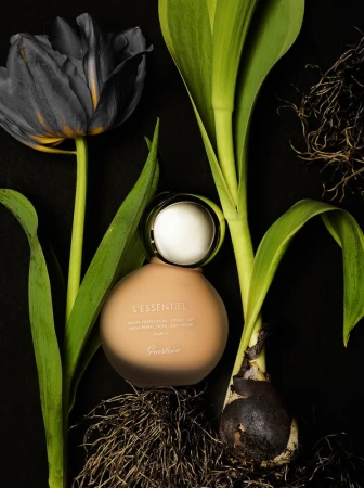 Still life shot of Guerlain L'Essential foundation bottle surrounded by black tulip and green plant stems against a black background.