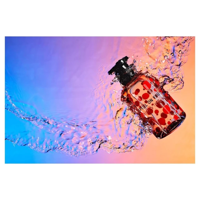 Product shot of a bottle of clear liquid with red polka dots floating in clear water with a gradient blue and peach background.