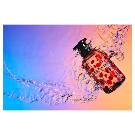Product shot of a bottle of clear liquid with red polka dots floating in clear water with a gradient blue and peach background.