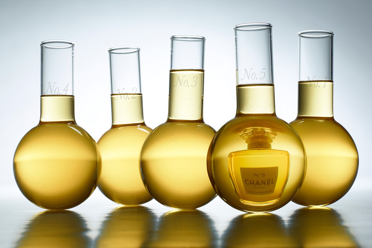 Laboratory glassware with numbered labels contains fragrance, with Chanel No. 5 bottle reflected in the liquid contents of one of the flasks.