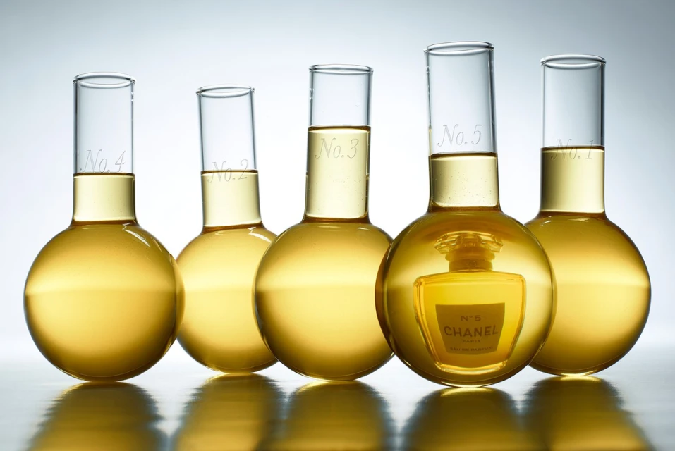 Laboratory glassware with numbered labels contains fragrance, with Chanel No. 5 bottle reflected in the liquid contents of one of the flasks.