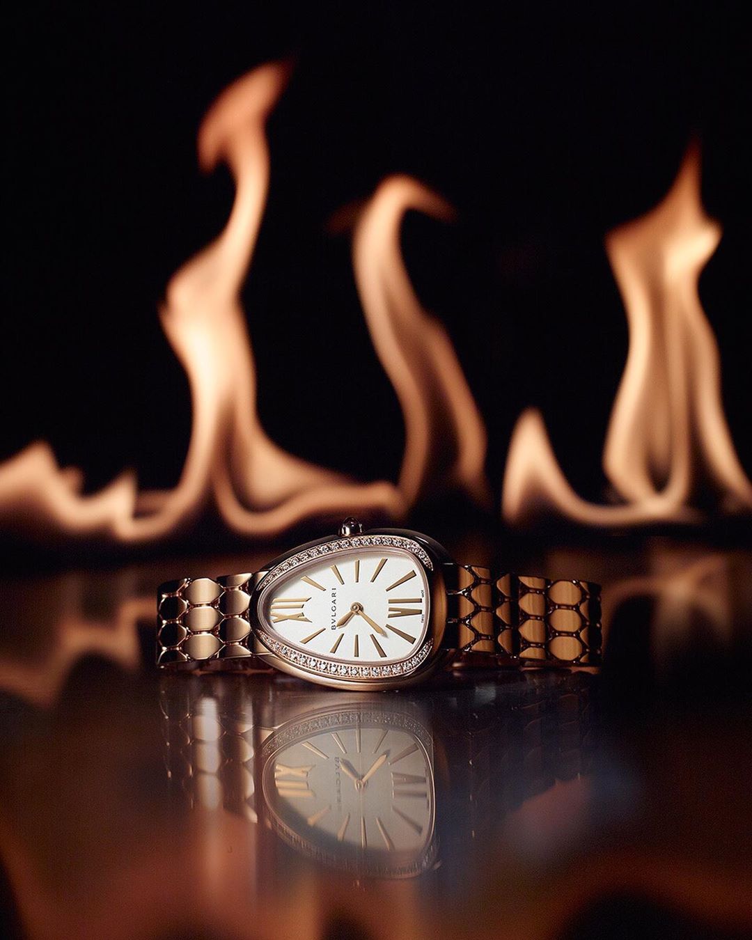 Close-up of a gold Bulgari Serpenti watch with diamonds, set against a dramatic black background with flames, casting a warm glow.