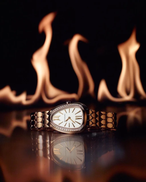 Close-up of a gold Bulgari Serpenti watch with diamonds, set against a dramatic black background with flames, casting a warm glow.