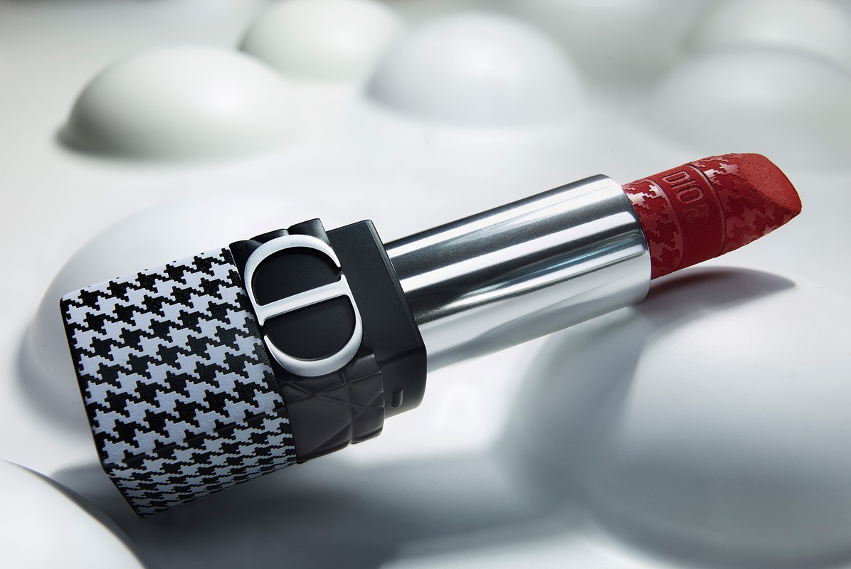 Close-up product photograph of a Dior lipstick with a houndstooth cap and debossed red bullet on a textured white surface.