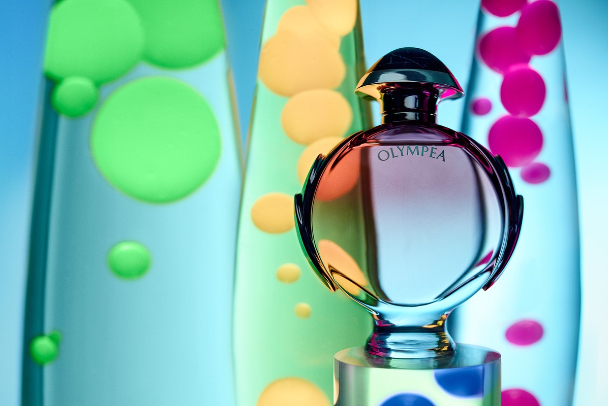 Perfume bottle of Olympea standing on a polka-dot platform with three lava lamps in the background, lit with pastel colors.