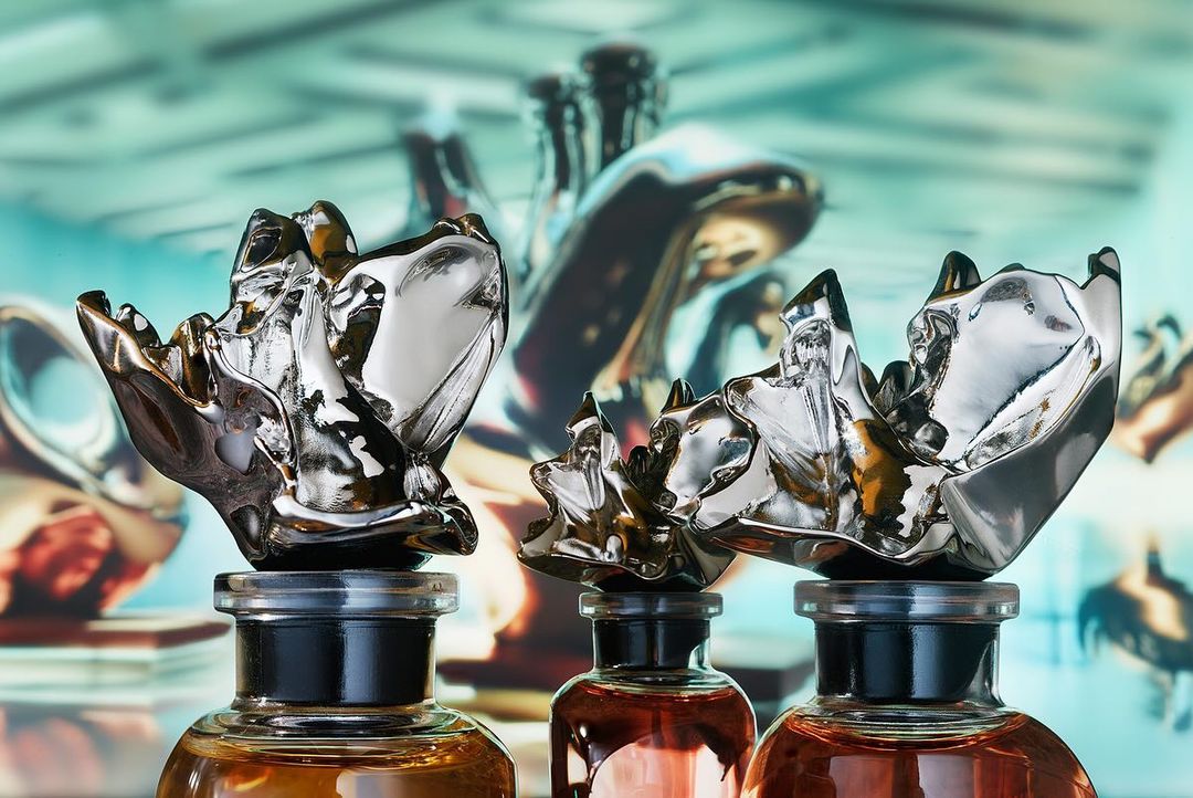 Three amber-colored glass perfume bottles are closely lined, each capped with an artistic silver stopper, set against an abstract backdrop.