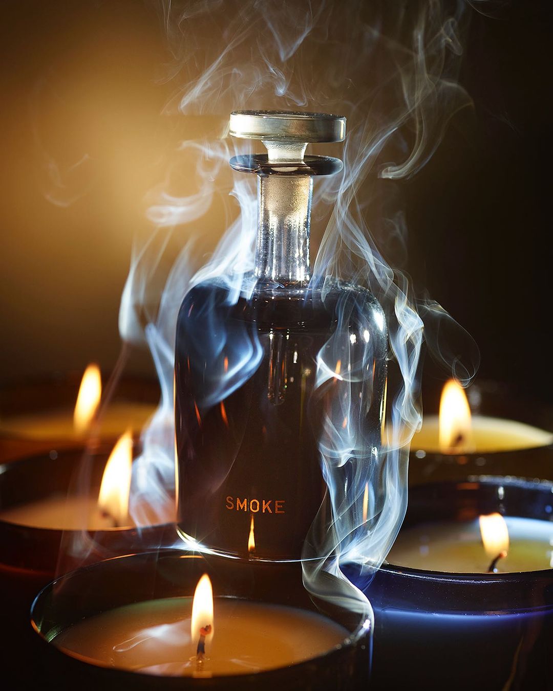 Elegant product shot of a glass bottle with golden accents surrounded by candles and swirling smoke against a dark gradient background.