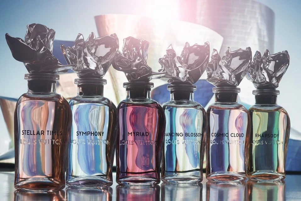 Row of Louis Vuitton perfume bottles with decorative toppers, displayed against a modern building backdrop on a reflective surface.