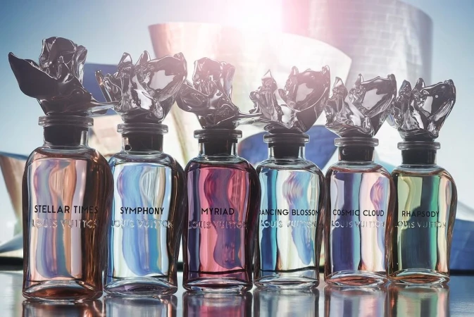 Row of Louis Vuitton perfume bottles with decorative toppers, displayed against a modern building backdrop on a reflective surface.