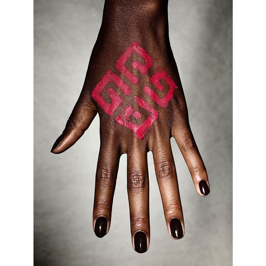 Close-up shot of a dark-skinned adult hand, featuring dark nail polish and a geometric red design painted on the back of the hand.