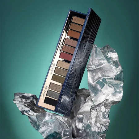 Eye shadow palette by Charlotte Tilbury on silver crinkled material against a teal background in a beauty product shot.