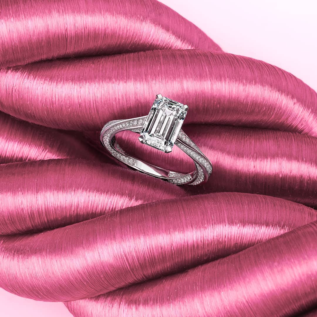 Close-up of a diamond ring with an emerald-cut stone on a twisted band, set against a backdrop of pink satin fabric.