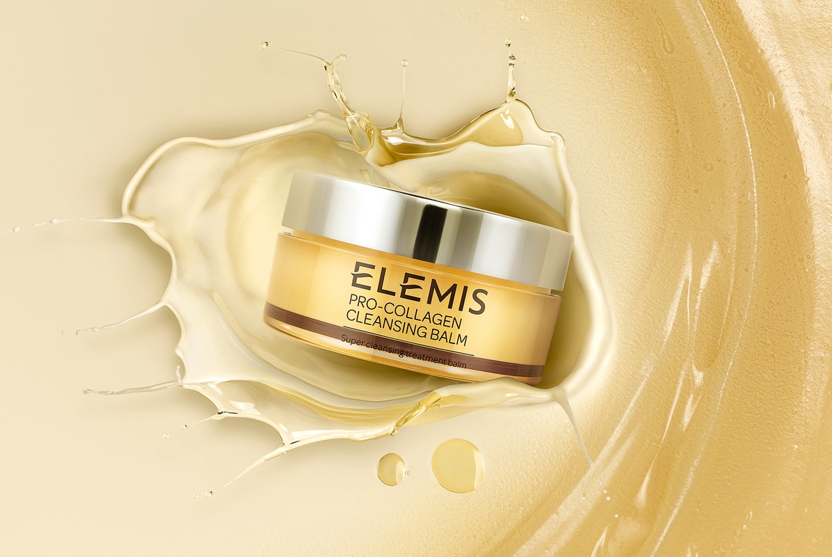 Elevated view of a gold-colored Elemis cleansing balm jar with a silver lid, surrounded by a splash of liquid against a tan backdrop.