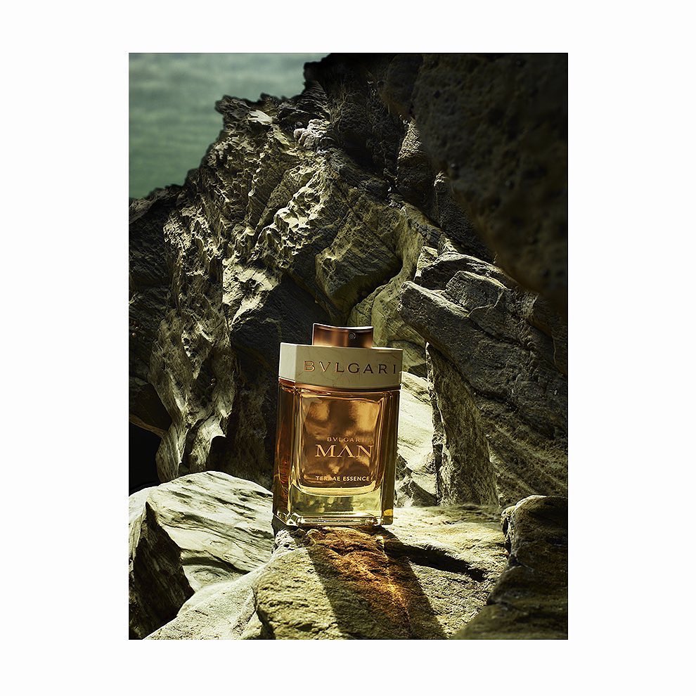 A rectangular glass fragrance bottle resting on dark weathered rocks outdoors, captured in a vertical composition.