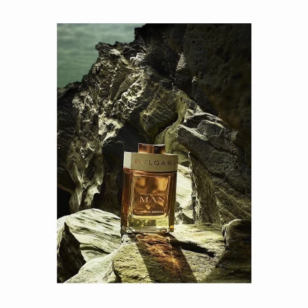 A rectangular glass fragrance bottle resting on dark weathered rocks outdoors, captured in a vertical composition.