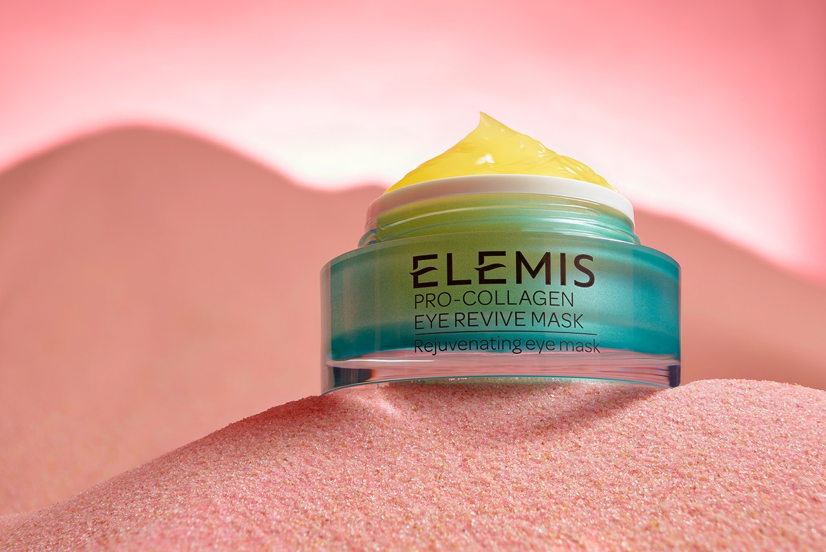 Elemis Pro-Collagen Eye Revive Mask jar with yellow cream on a pink sandy surface against a soft pink background, skincare product.