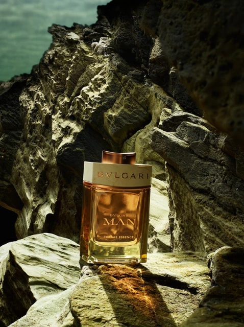 BVLGARI MAN Terrae Essence fragrance bottle prominently displayed on textured rock formations, set against a soft, blurred coastal background.