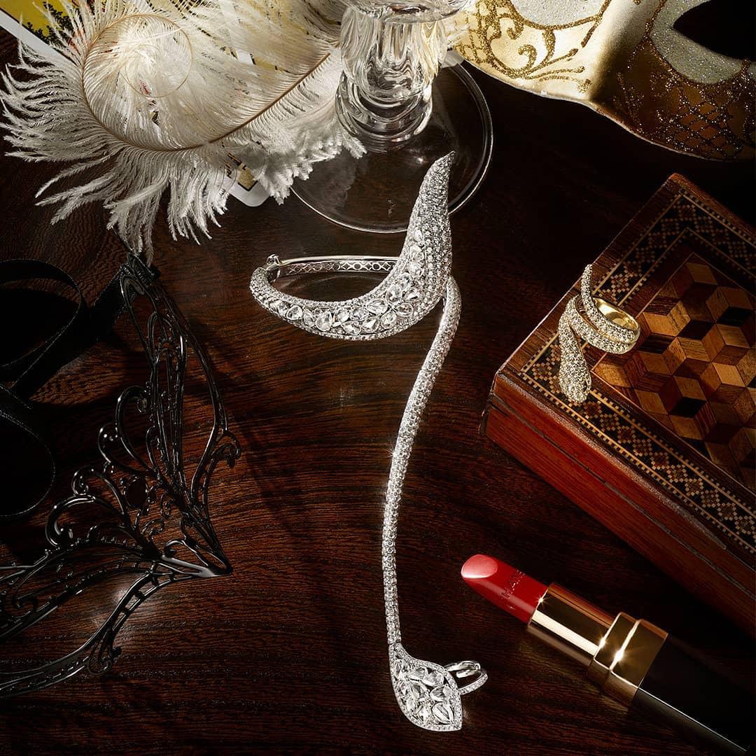 Still life composition featuring a diamond bracelet and ring displayed near a black feathered mask, lipstick, glass, and decorative box.