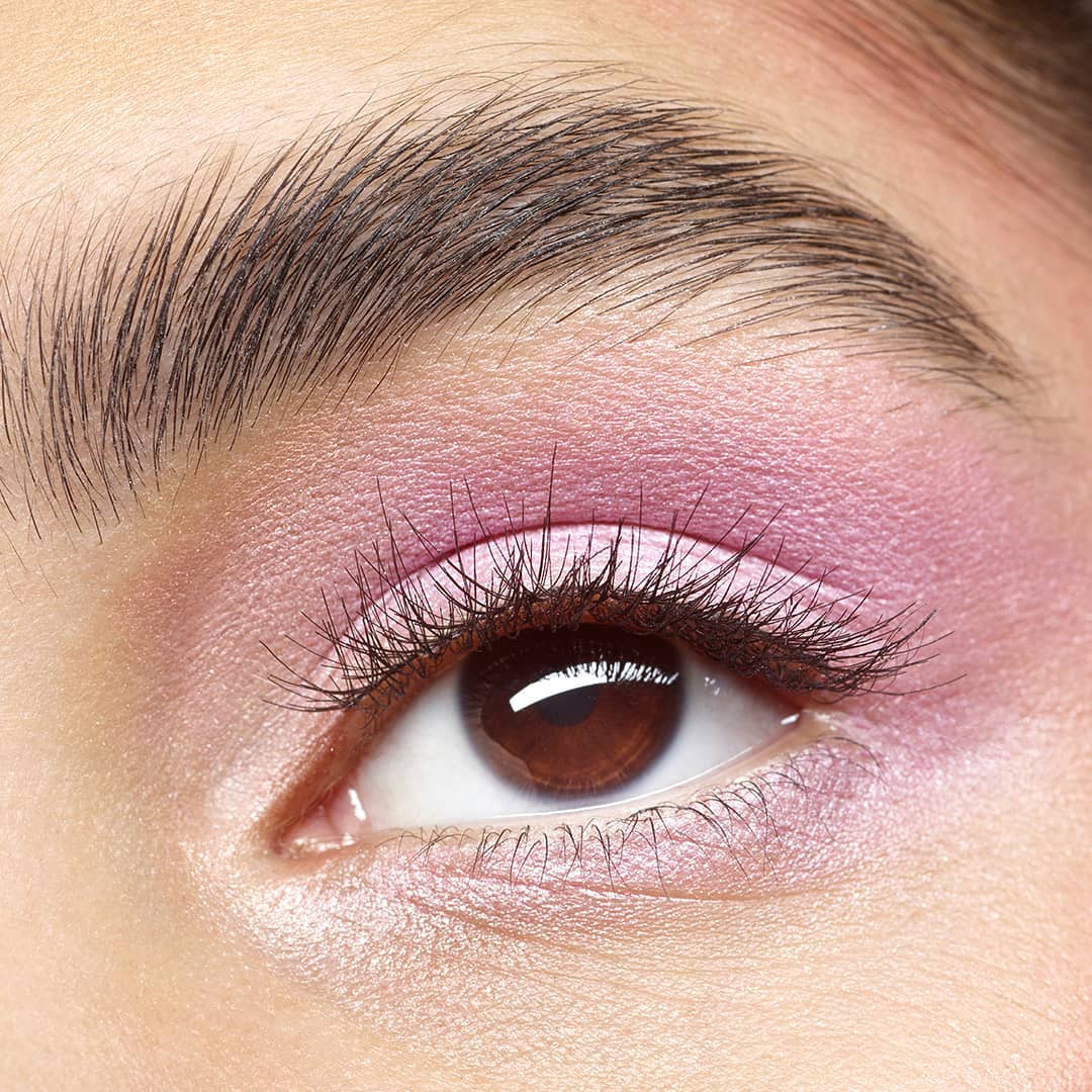 Close-up of a woman's eye with pink eyeshadow and dark brown eyebrow, eyelashes, and eye color against fair skin.