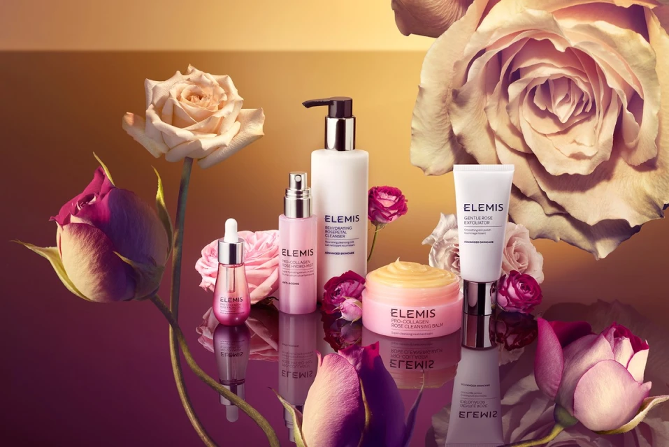 Elevated still life shot of Elemis skincare products including cleanser, balm, and exfoliator among rose blossoms on a reflective surface.