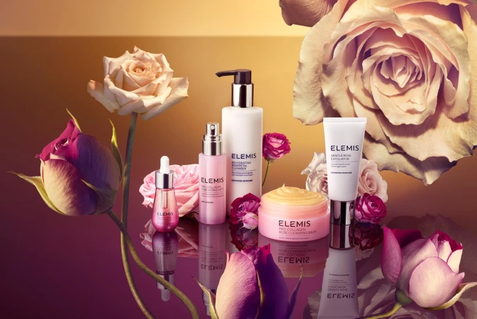 Elevated still life shot of Elemis skincare products including cleanser, balm, and exfoliator among rose blossoms on a reflective surface.