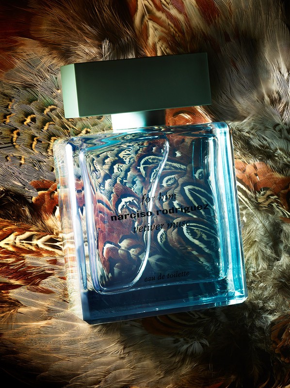 Close-up of a rectangular Narciso Rodriguez perfume bottle with a light green cap set against a feathered background in a studio shot.