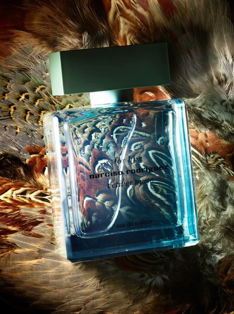 Close-up of a rectangular Narciso Rodriguez perfume bottle with a light green cap set against a feathered background in a studio shot.