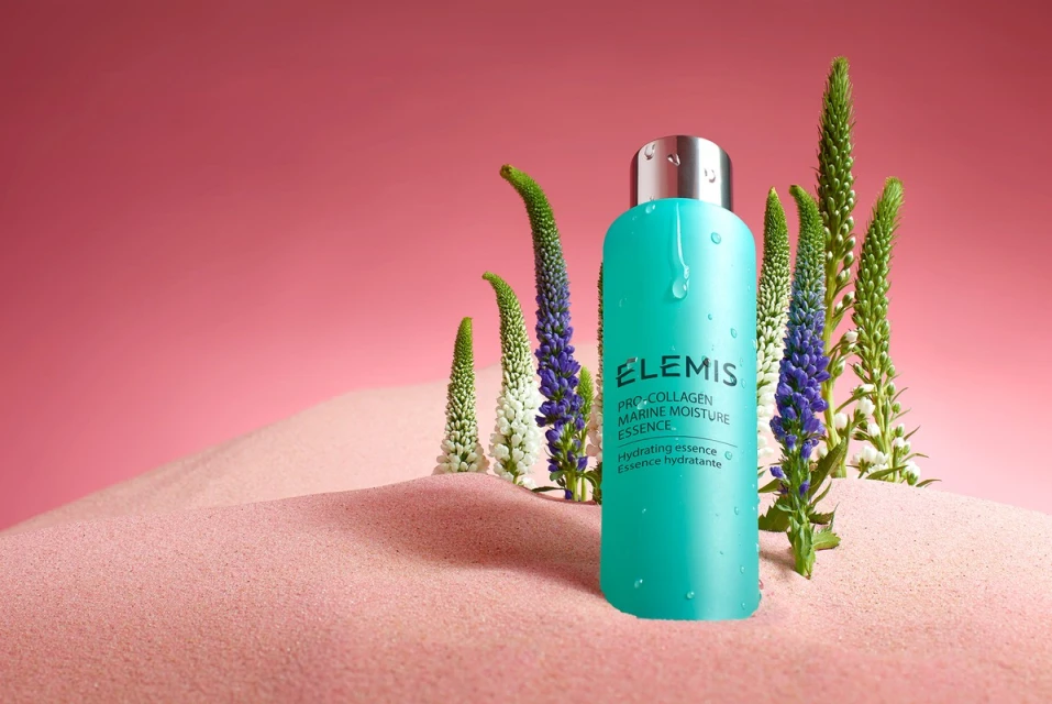 Teal ELEMIS Pro-Collagen Marine Moisture Essence bottle sits in a pink sand dune surrounded by flowering plants against a pink backdrop.