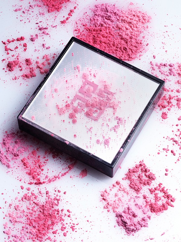 Close-up product shot of a makeup compact case with a mirror and pink blush powder around and on the cosmetic case on a white surface.
