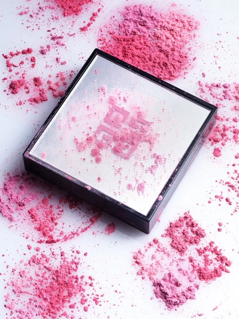 Close-up product shot of a makeup compact case with a mirror and pink blush powder around and on the cosmetic case on a white surface.