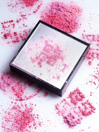 Close-up product shot of a makeup compact case with a mirror and pink blush powder around and on the cosmetic case on a white surface.