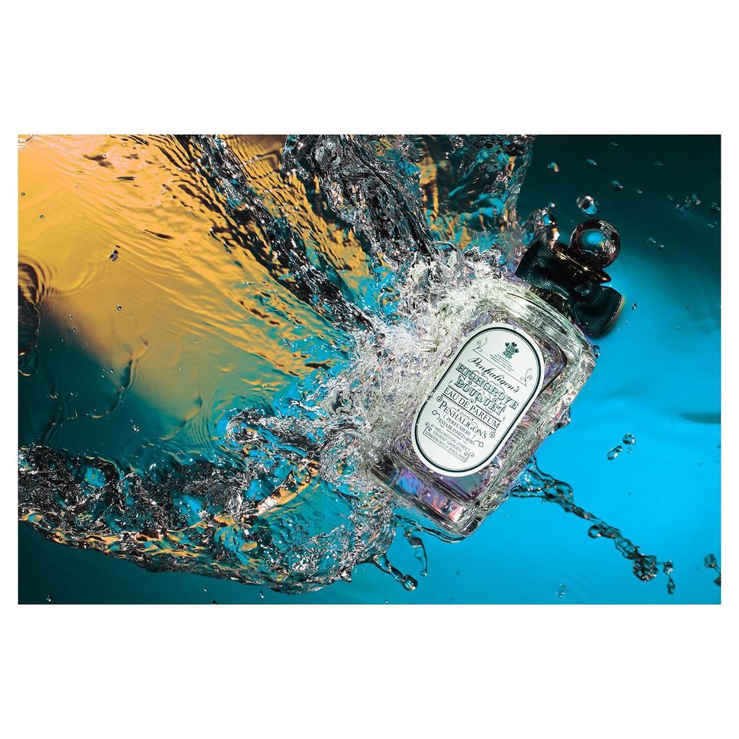 A perfume bottle submerged in water surrounded by dynamic splashes against a blue and yellow gradient background.