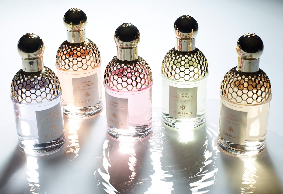 Five perfume bottles from Guerlain's Aqua Allegoria line arranged on a reflective white surface, showcasing their golden honeycomb tops.