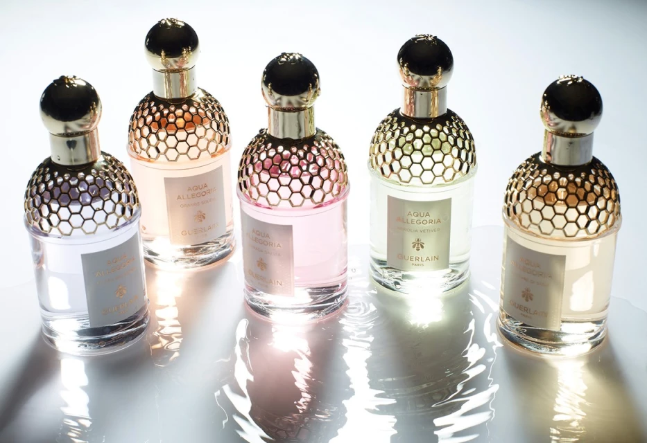Five perfume bottles from Guerlain's Aqua Allegoria line arranged on a reflective white surface, showcasing their golden honeycomb tops.