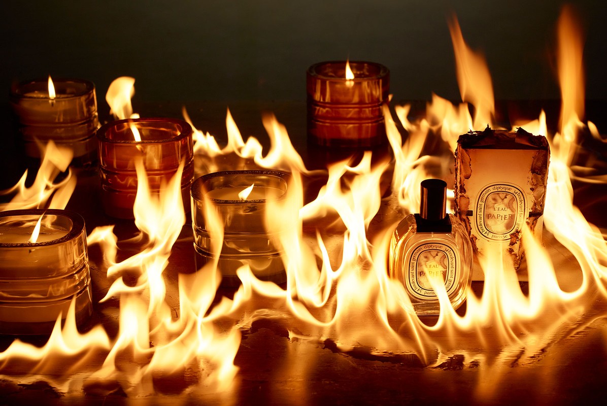Burning candles and perfume bottles on a wood surface with flames licking up around the glass in a warm, fiery tableau.