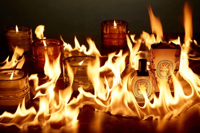 Burning candles and perfume bottles on a wood surface with flames licking up around the glass in a warm, fiery tableau.