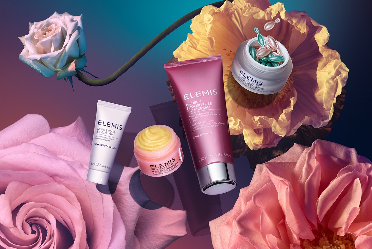 Elevated shot of ELEMIS skincare products including body cream and capsules nestled amid roses and flowers, showcasing beauty and skincare.