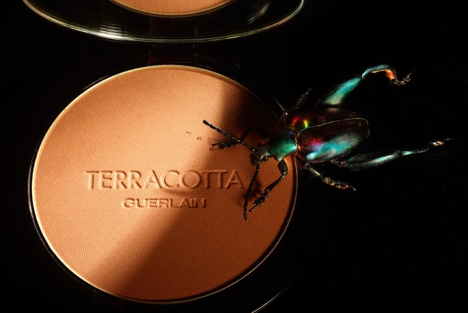Close-up product shot: Guerlain bronzer with a colorful beetle crawling on the makeup's surface, set against a stark black backdrop.