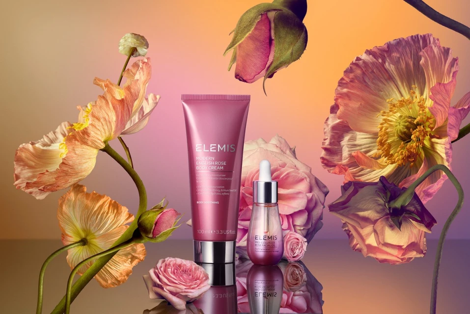 Cosmetic products, Elemis Modern English Rose body cream and facial oil surrounded by pink rose blossoms and buds, on a reflective surface.