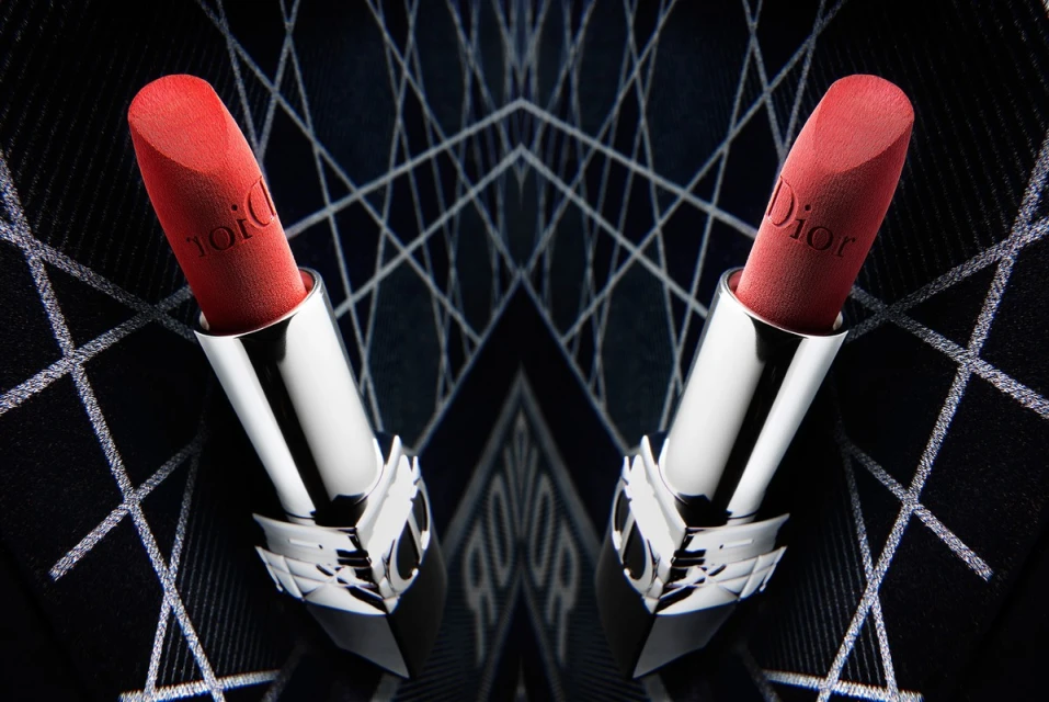 Close-up studio shot of two red Dior lipsticks with silver tubes symmetrically arranged against a dark geometric background.