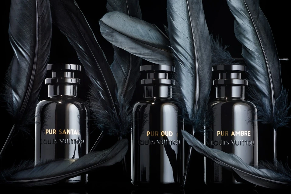 Three dark perfume bottles from Louis Vuitton are displayed with dark feathers against a black background, creating a moody, elegant aesthetic.