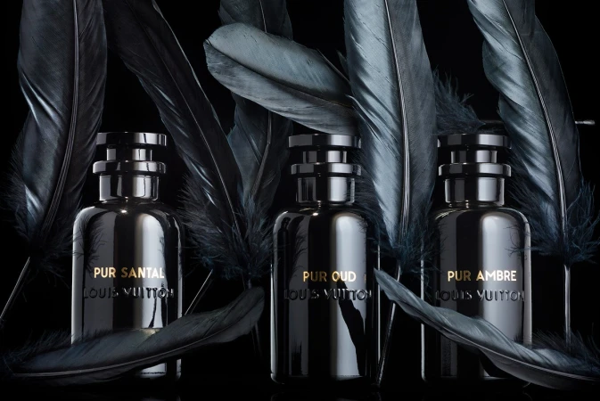Three dark perfume bottles from Louis Vuitton are displayed with dark feathers against a black background, creating a moody, elegant aesthetic.