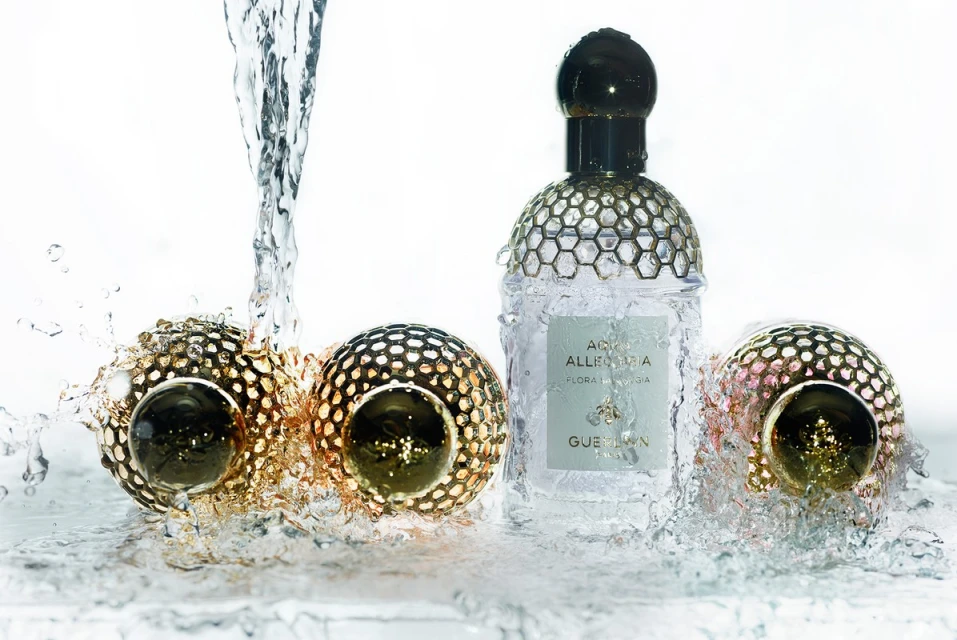 Perfume bottle and golden ornaments with water splashing over them on a white surface, product shot for beauty and cosmetics.