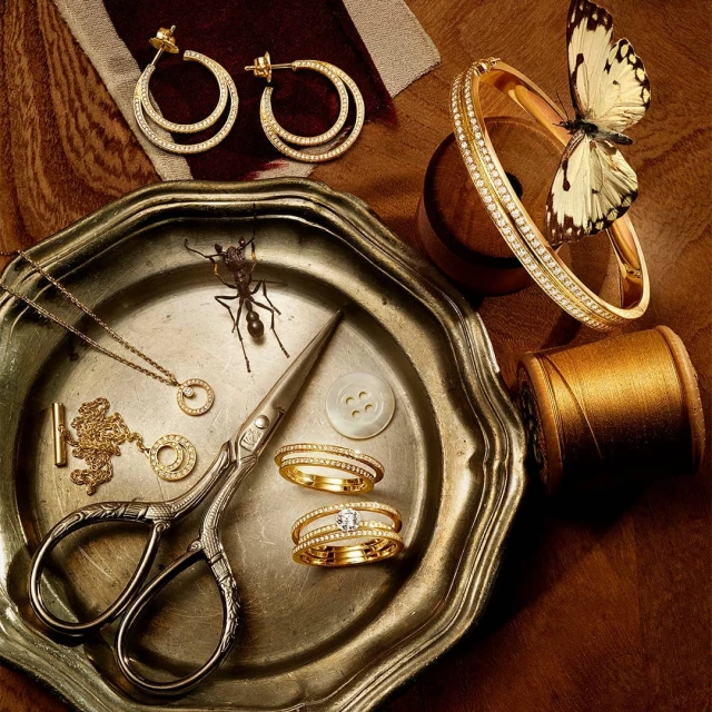 Close-up of gold jewelry, including earrings, rings, necklace, and bracelet, arranged on a silver tray with button, thread, and butterfly.