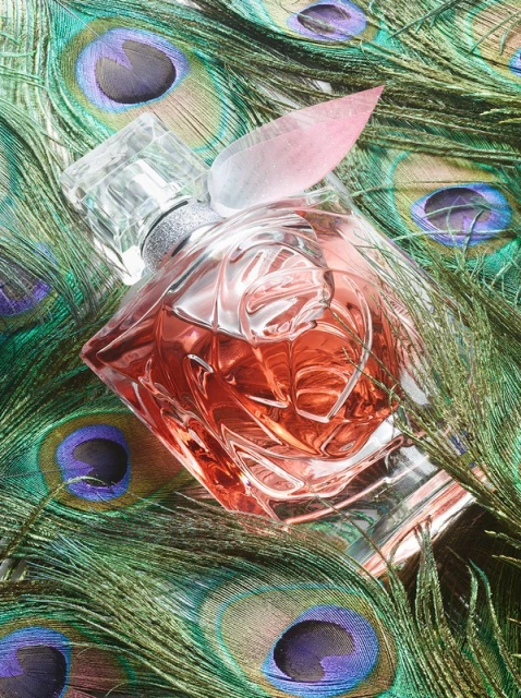 Elegant still life featuring a crystal perfume bottle with a pink tint, resting on a bed of iridescent peacock feathers. Beauty product.