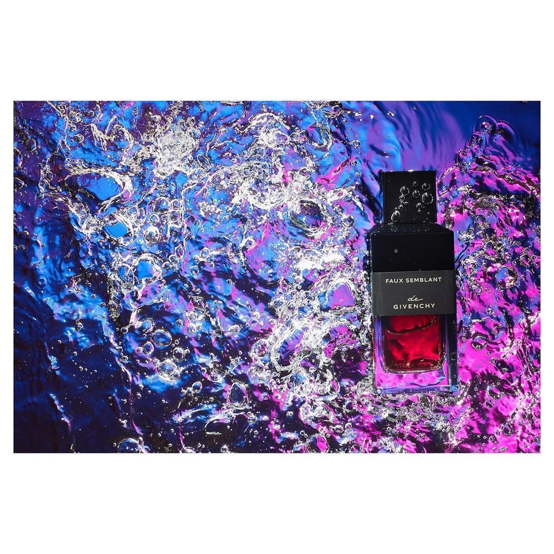 A black perfume bottle with a red liquid inside is set against a vibrant background of swirling blue, purple, and white water.